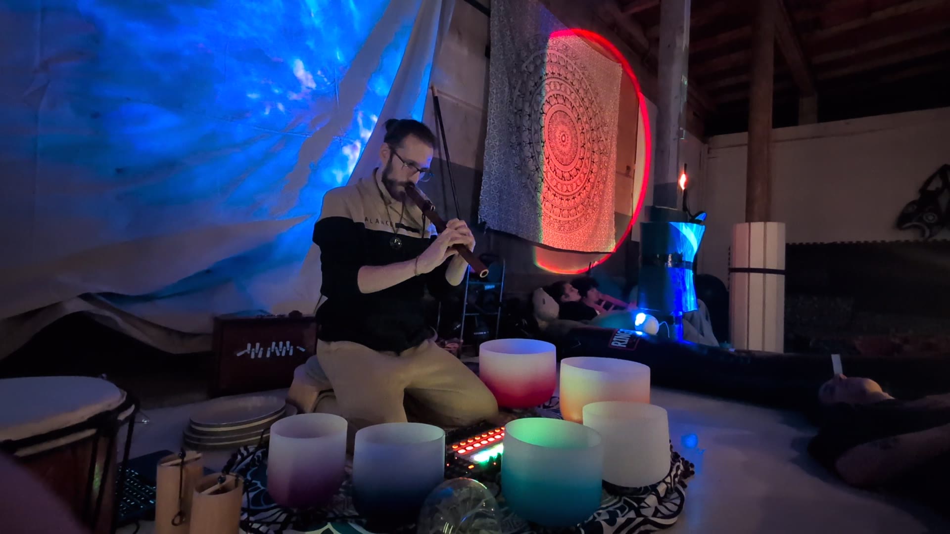Ananda playing a flute behind a ring of crystal bowls, people lying down behind him